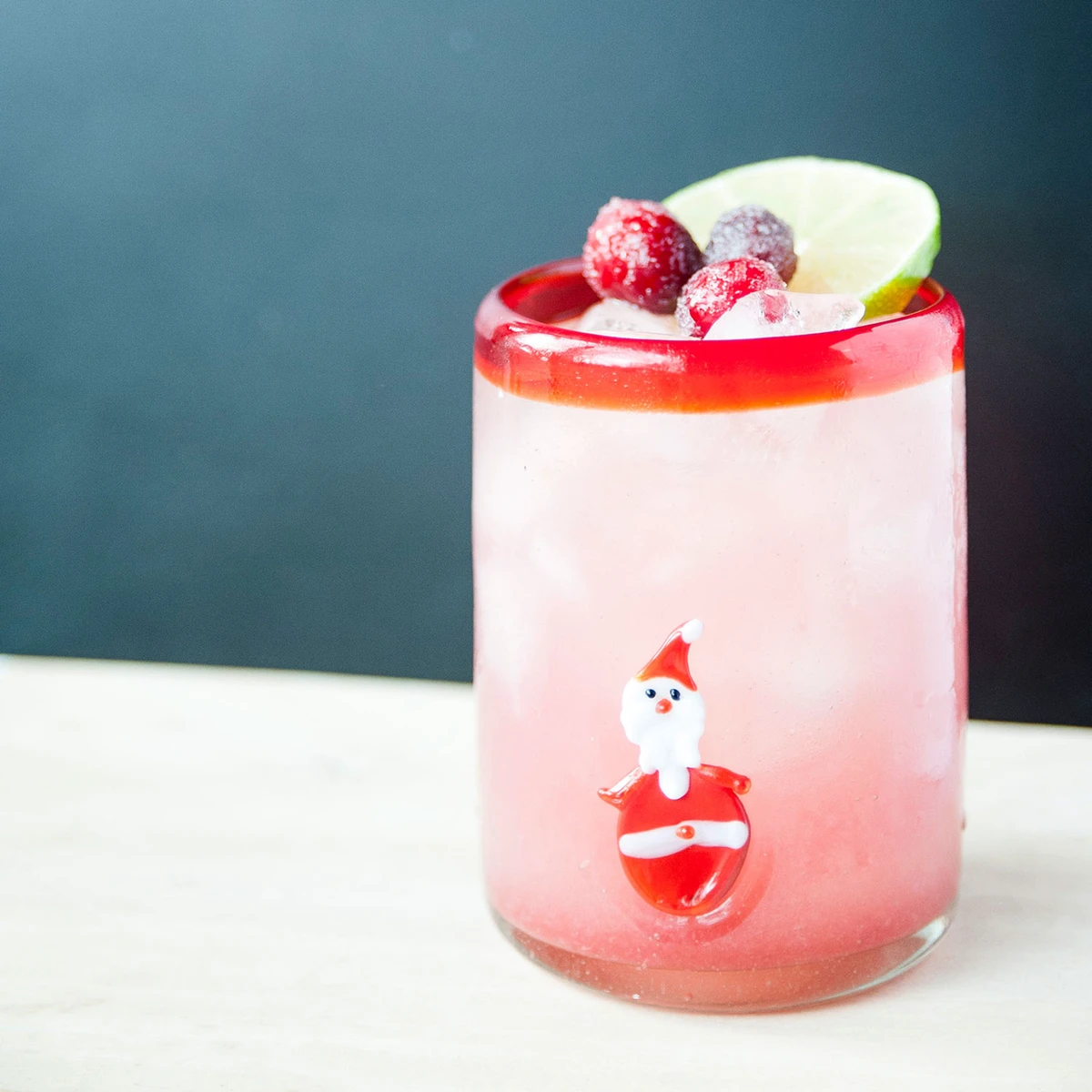A cocktail class with a santa on the side, filled with a pink liquid and topped with lime and cranberries