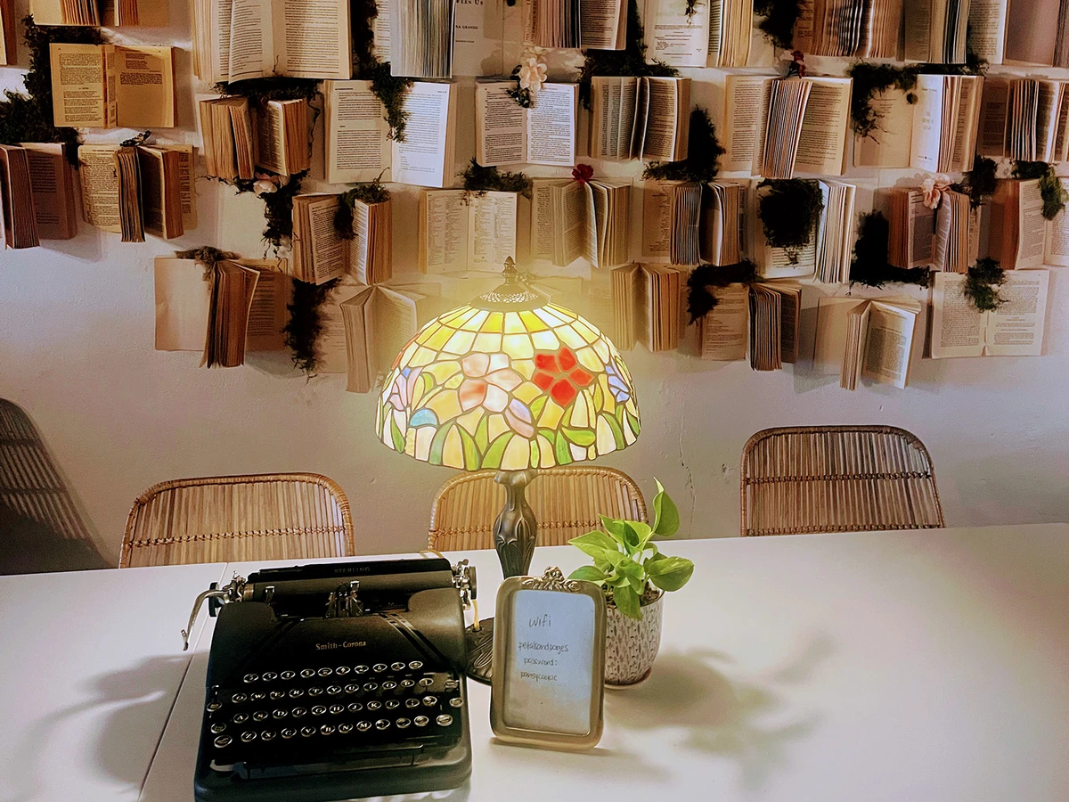 Typewriter and an antique lamp sit on a desk in front of a wall covered in open books