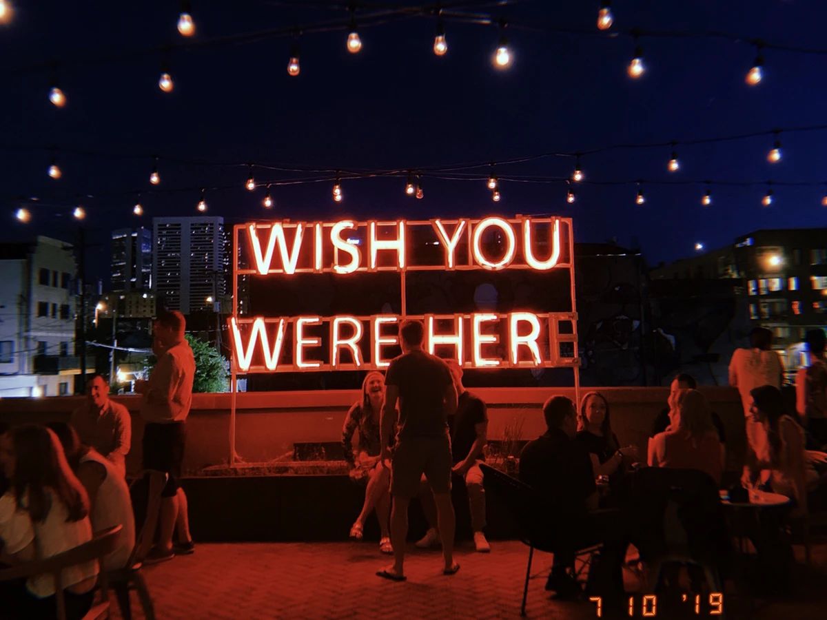 People sit and chat in front of the "Wish You Were Here" neon sign