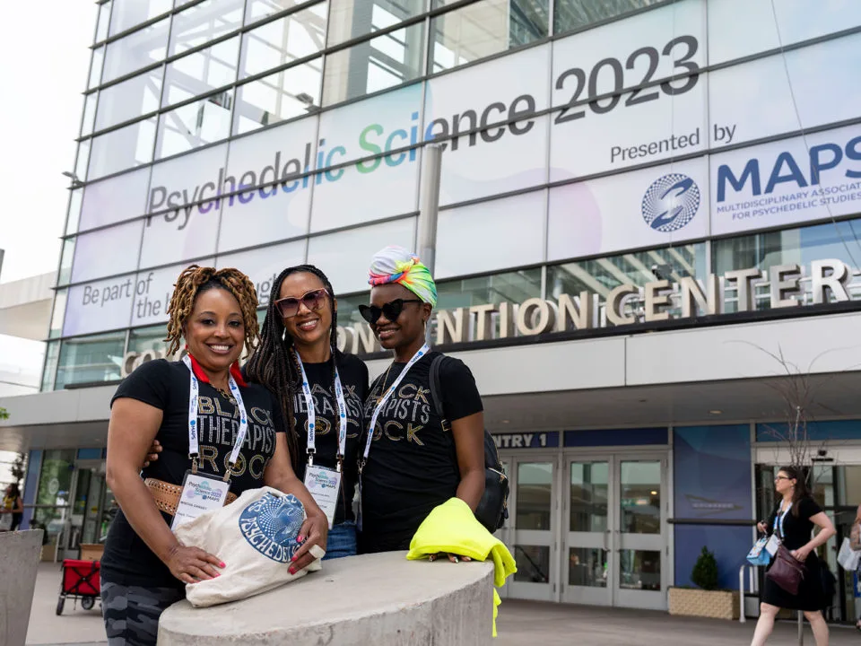 Three black women smile in front of the Psychedelic Science 2023 conference banner