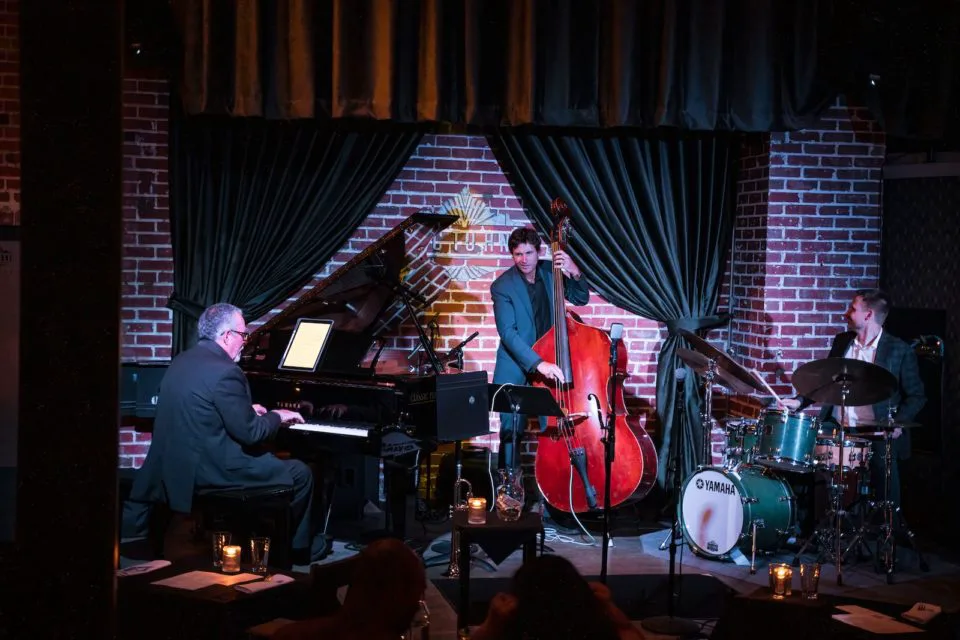 A piano player, a stand-up bass player, and a drummer on a stage at a dimply lit jazz club with a black curtain and brick wall in the background