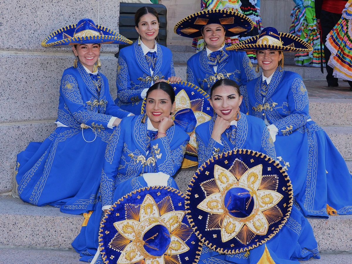 Six women dressed in traditional Mexican dresses pose on steps
