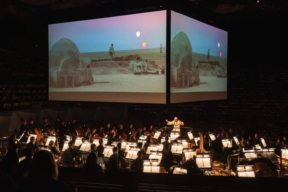 The orchestra performs under a giant screen playing a movie scene.