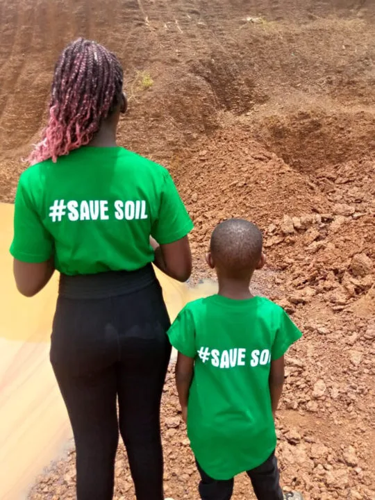Woman and child stand in front of a hill of dirt wearing shirts that save #savesoil