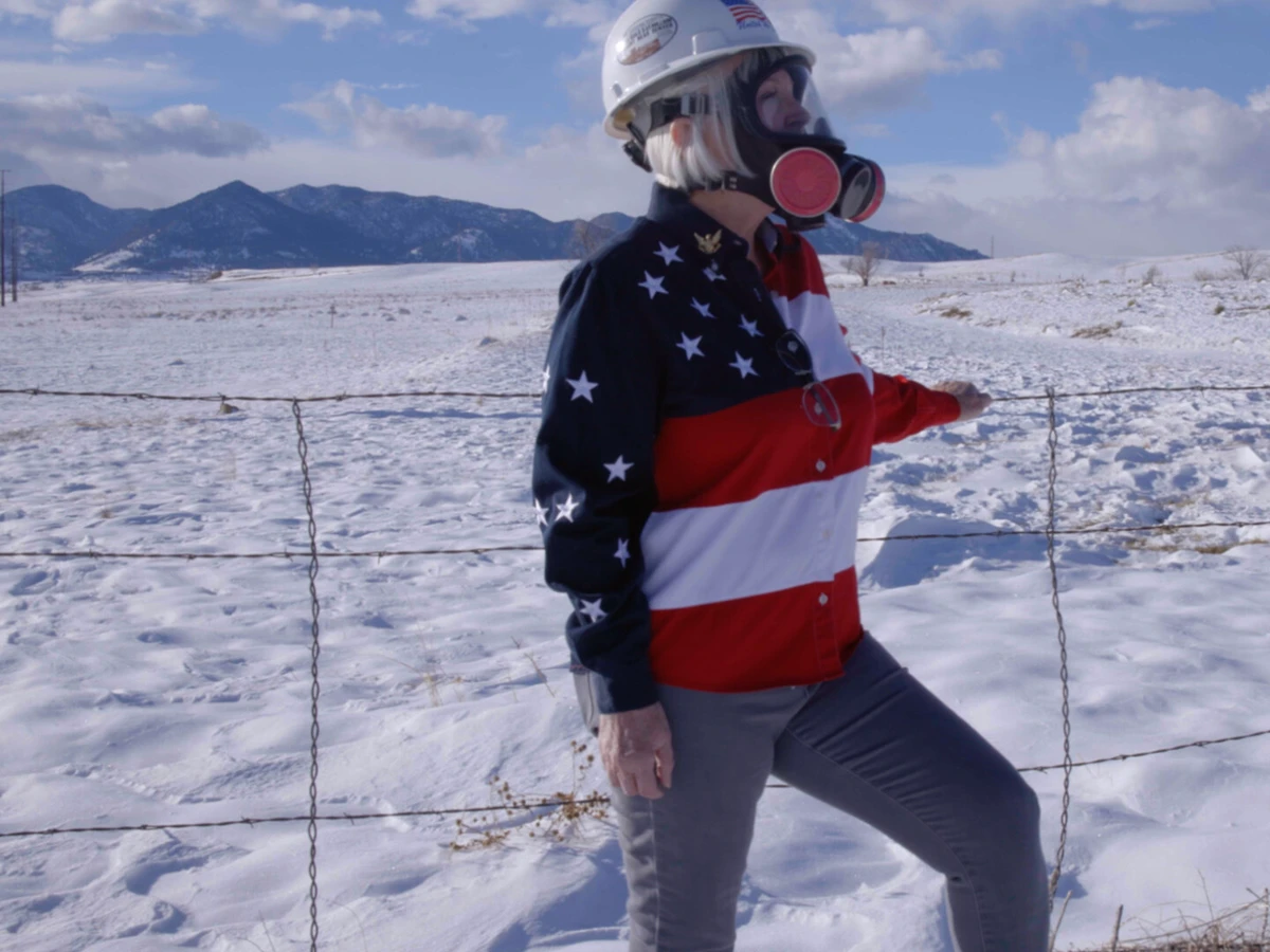 Woman in an American flag shirt stands in front of a field wearing a gas mask