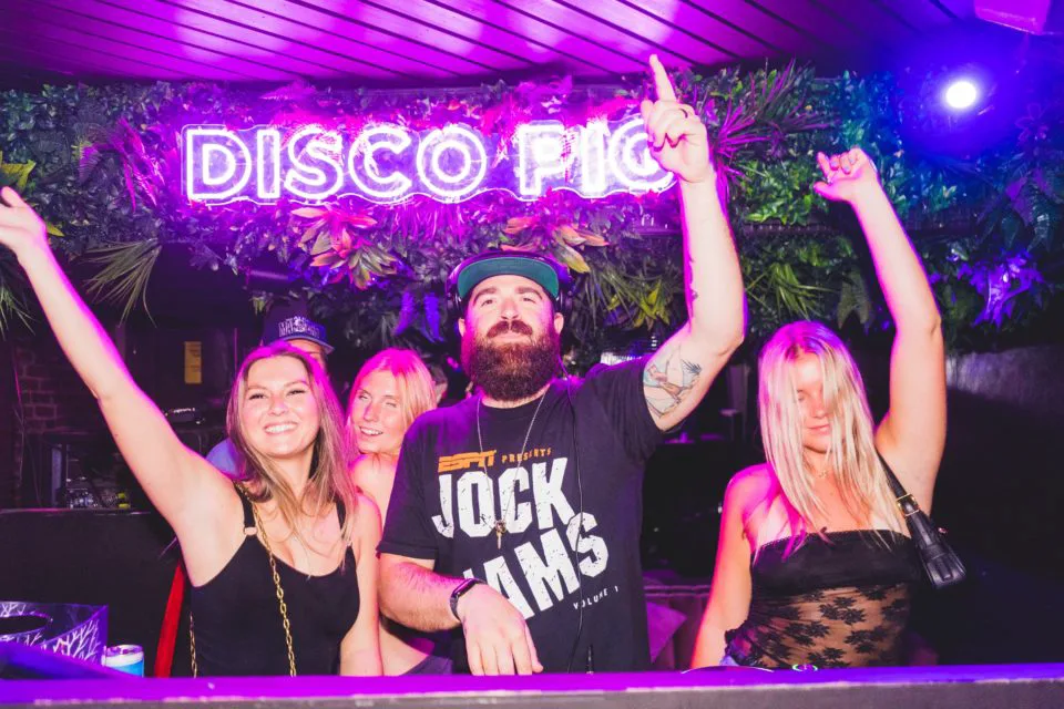 Three people dance at the DJ booth with a neon sign in the back reading "Disco Pig"