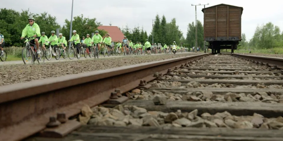 A still from For The Living where cyclists pedal next to tracks leading to Auschwitz-Birkenau