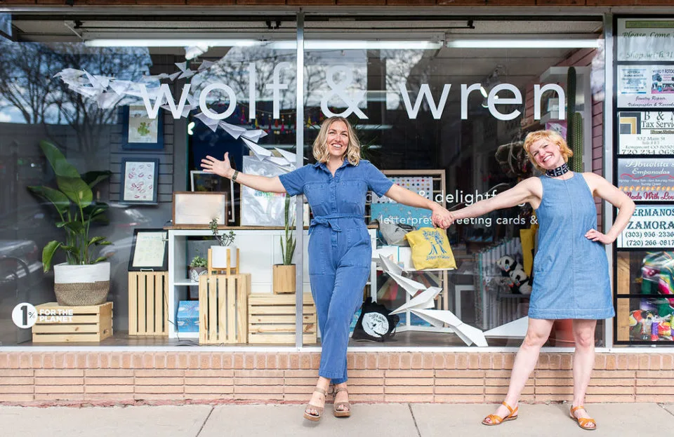 Lauren Stapleton and Liz Wolf stand in front of Wolf & Wren's storefront, holding hands and smiling.