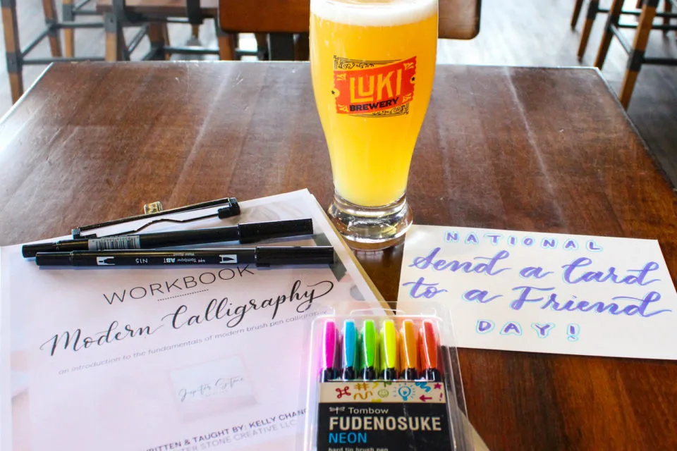 A workbook about calligraphy, pens, and a card that reads "Send a Card to a Friend Day!" in front of a glass of beer.