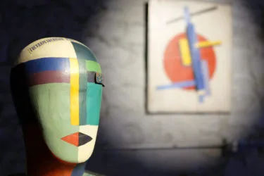 A facelike abstract sculpture sits in front of an abstract painting of red, yellow, and blue.
