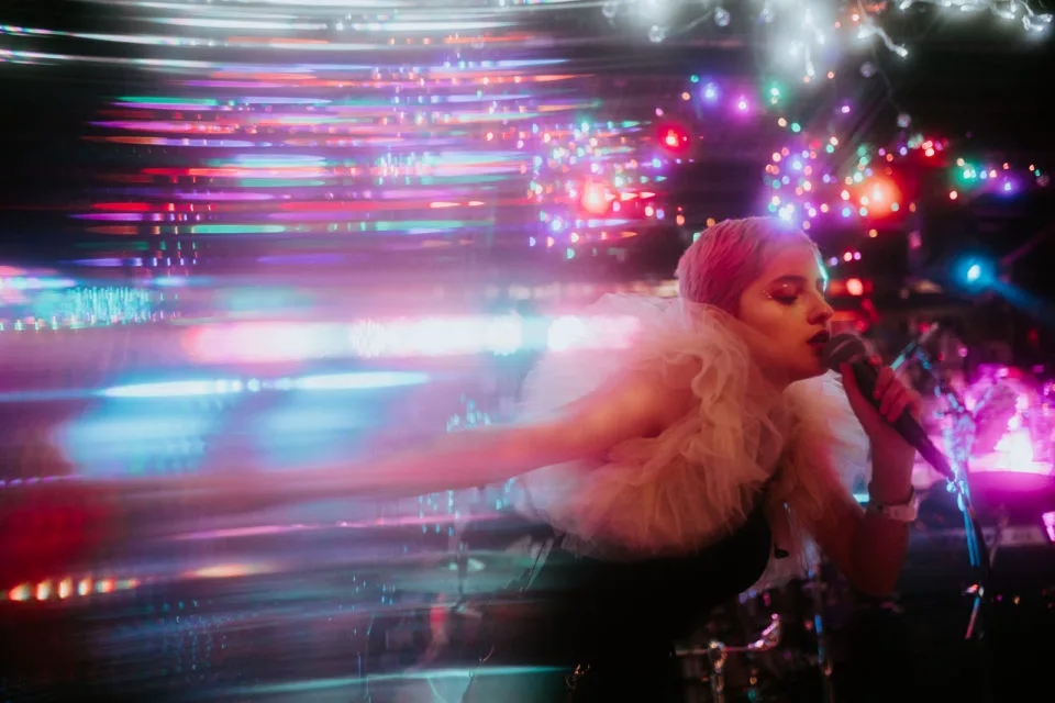 Denver singer Neoma sings into a handheld microphone against a bright, colorful wall of lights